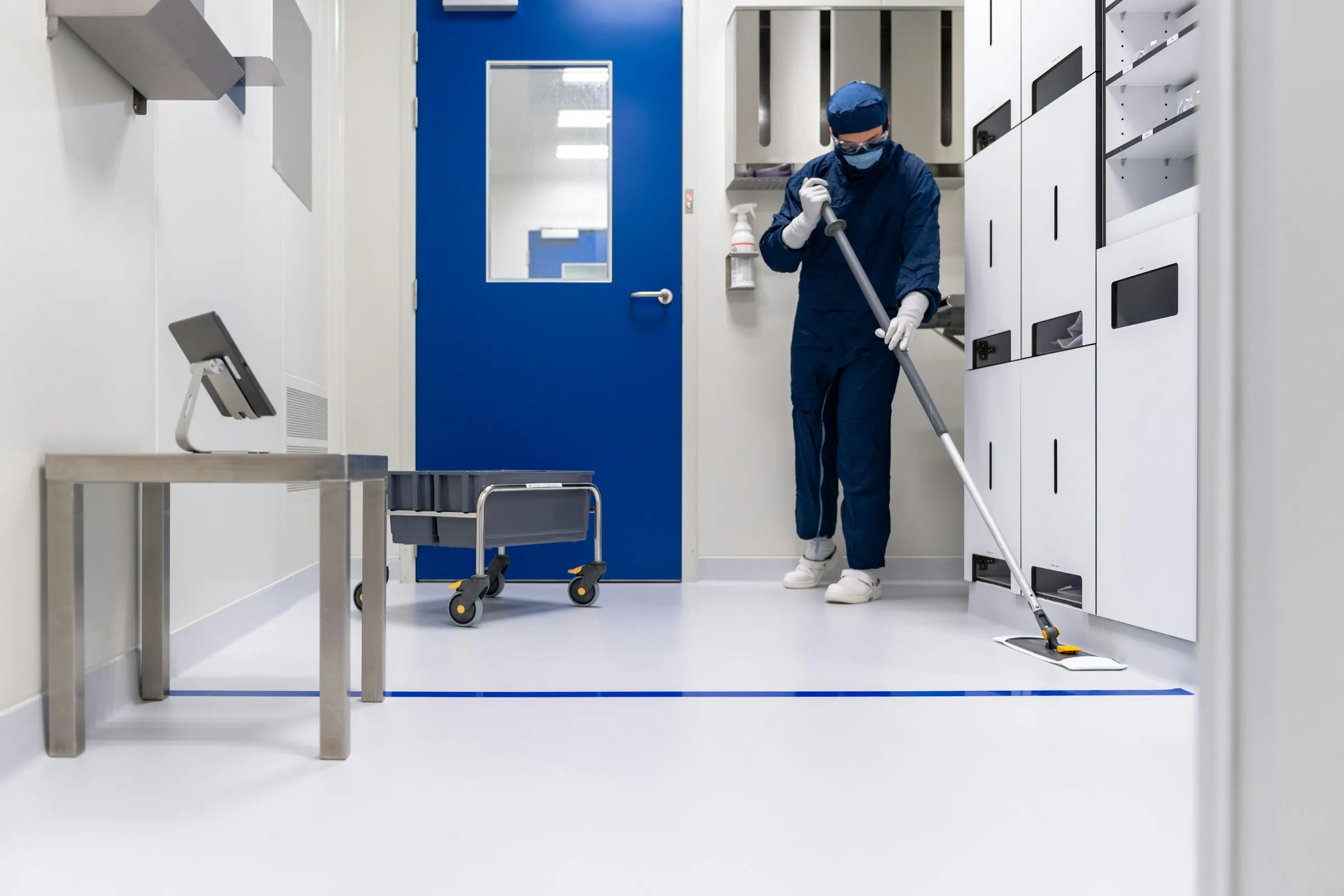 A cleaner in protective clothing mopping a bright clinical workspace.